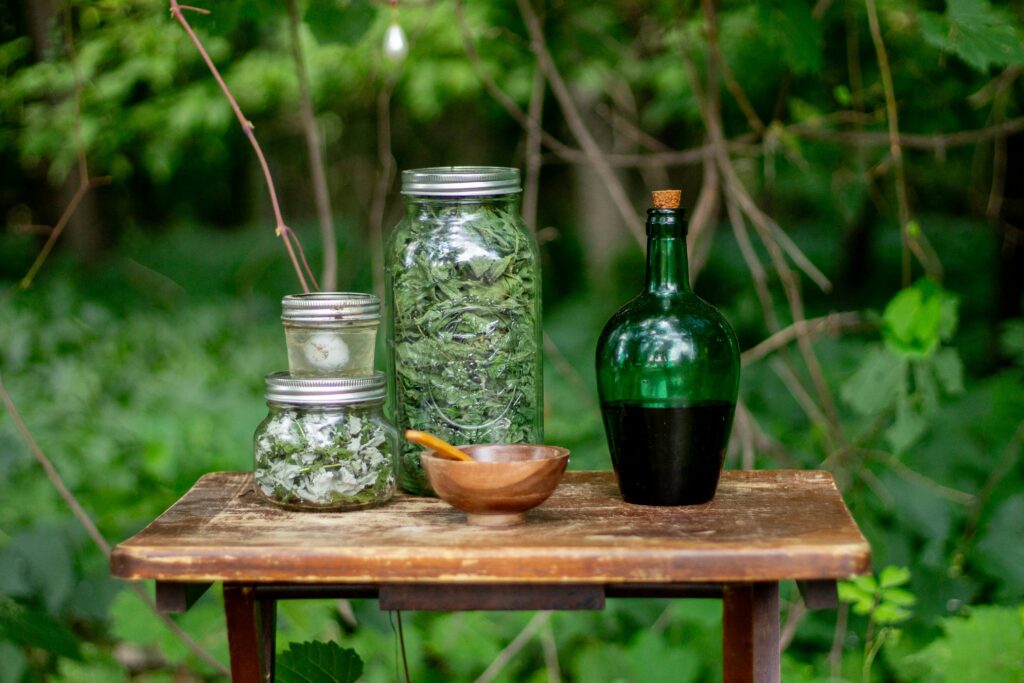 Glass jars of dried herbs and a green bottle on a wooden table outdoors.